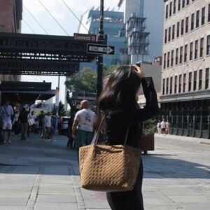 May include: A brown woven tote bag with a shoulder strap is carried on a city street. The bag has a textured pattern and a rectangular shape. The person is wearing a black dress and boots. The street sign reads "Greenwich St."