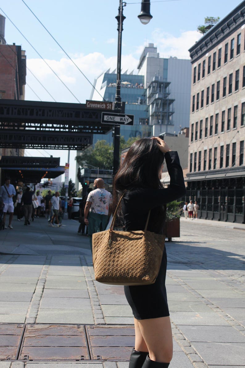 May include: A brown woven tote bag with a shoulder strap is carried on a city street. The bag has a textured pattern and a rectangular shape. The person is wearing a black dress and boots. The street sign reads "Greenwich St."