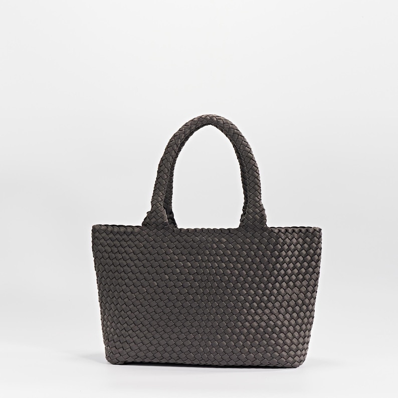 May include: A dark gray tote bag with a woven design. The bag has two braided handles and a rectangular shape. The material appears to be a soft, flexible fabric. The bag is set against a plain white background.