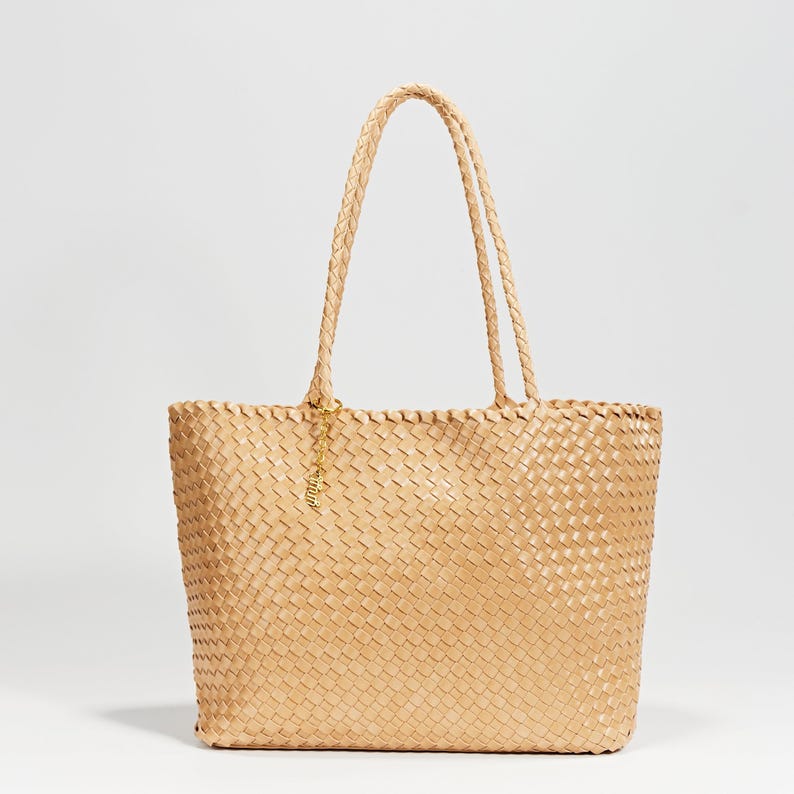 Pode incluir: Uma bolsa tote bege tecida com al&ccedil;as tran&ccedil;adas e um pingente dourado. A bolsa apresenta um formato retangular com um padr&atilde;o tecido por todo o corpo e al&ccedil;as. A bolsa foi projetada para transportar itens.