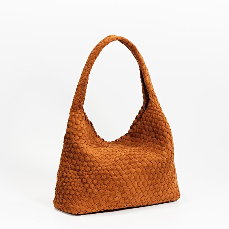K&ouml;nnte beinhalten: Eine braune Schultertasche mit geflochtenem Griff. Die Tasche hat eine Korbwebstruktur und eine halbmondf&ouml;rmige Form, die an der Basis breiter ist und sich nach oben verj&uuml;ngt. Warme, erdige braune Farbe.
