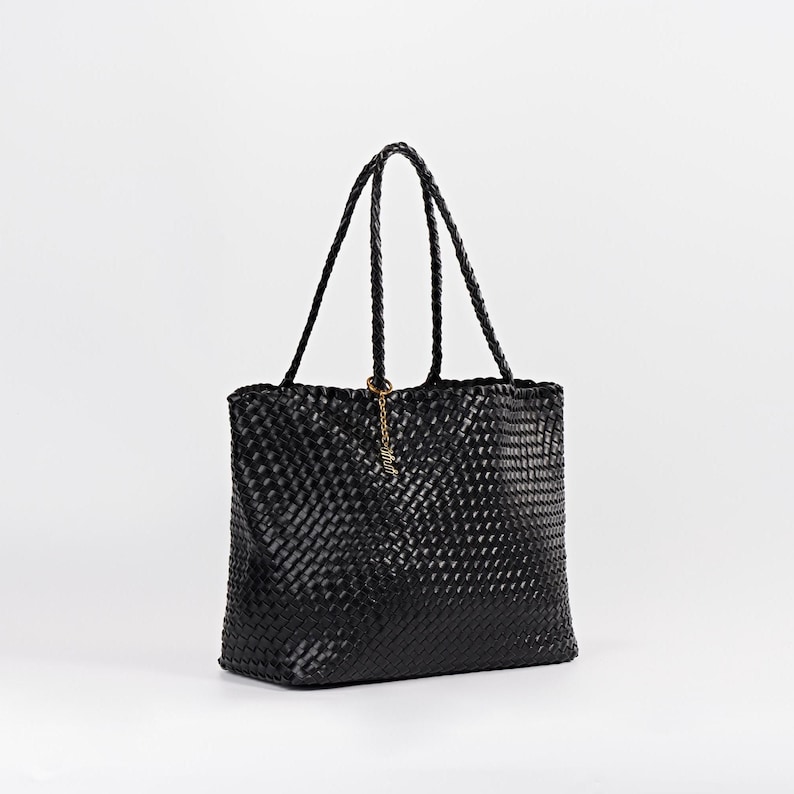 Pode incluir: Bolsa tote preta tecida com al&ccedil;as tran&ccedil;adas. A bolsa tem formato retangular e um pingente dourado pendurado em uma das al&ccedil;as. O padr&atilde;o de tecido &eacute; vis&iacute;vel em todo o exterior da bolsa.