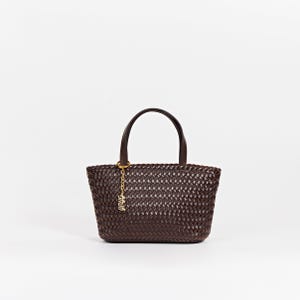 May include: A dark brown woven tote bag with two short handles and a gold-colored charm. The bag has a basket-weave design and a structured shape. The handles are a darker shade of brown.