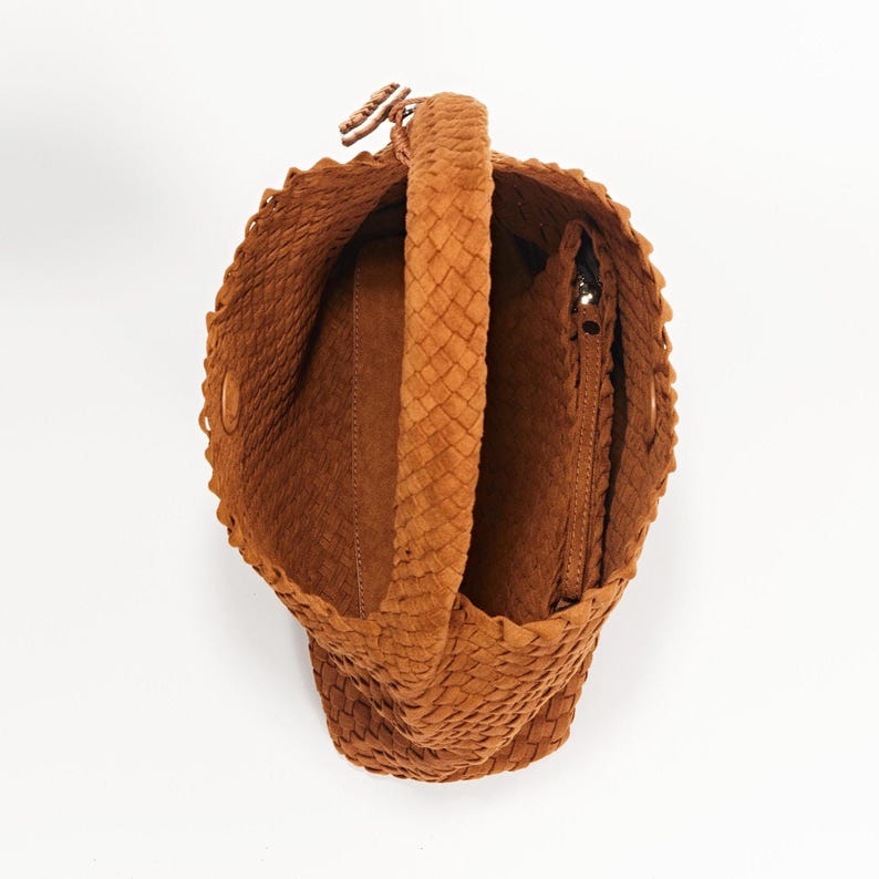 K&ouml;nnte beinhalten: Eine gewebte braune Handtasche mit geflochtenem Griff. Die Tasche hat eine runde Form und eine Rei&szlig;verschlusstasche im Inneren. Der Innenraum ist in einem helleren Braunton gehalten. Die Tasche ist aus weichem, strukturiertem Material gefertigt.