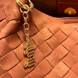 May include: Close-up of a brown woven handbag with a gold-colored keychain. The keychain has a gold ring, chain, and a cursive word charm. The handbag has a brown leather handle and a magnetic clasp.