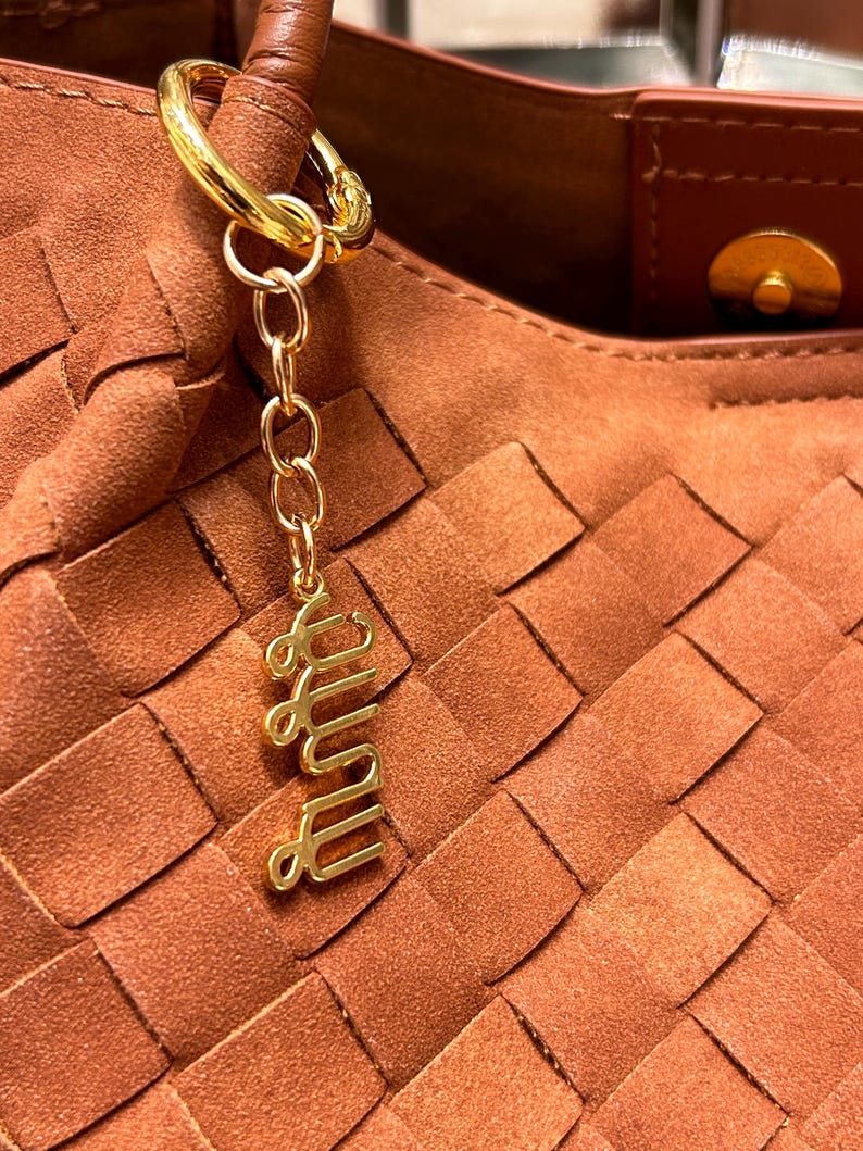 May include: Close-up of a brown woven handbag with a gold-colored keychain. The keychain has a gold ring, chain, and a cursive word charm. The handbag has a brown leather handle and a magnetic clasp.
