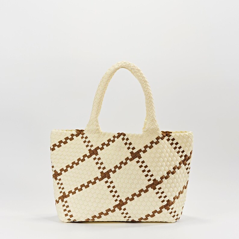 May include: Cream-colored tote bag with a woven design. The bag features a repeating pattern of brown squares and a braided handle. The bag is rectangular with a slightly rounded bottom.