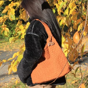 K&ouml;nnte beinhalten: Eine orangefarbene, gewebte Umh&auml;ngetasche mit einem dekorativen Anh&auml;nger. Die Tasche wird &uuml;ber einer schwarzen, flauschigen Jacke getragen. Der Hintergrund zeigt gelbe und gr&uuml;ne Bl&auml;tter, was auf eine herbstliche Umgebung hindeutet.