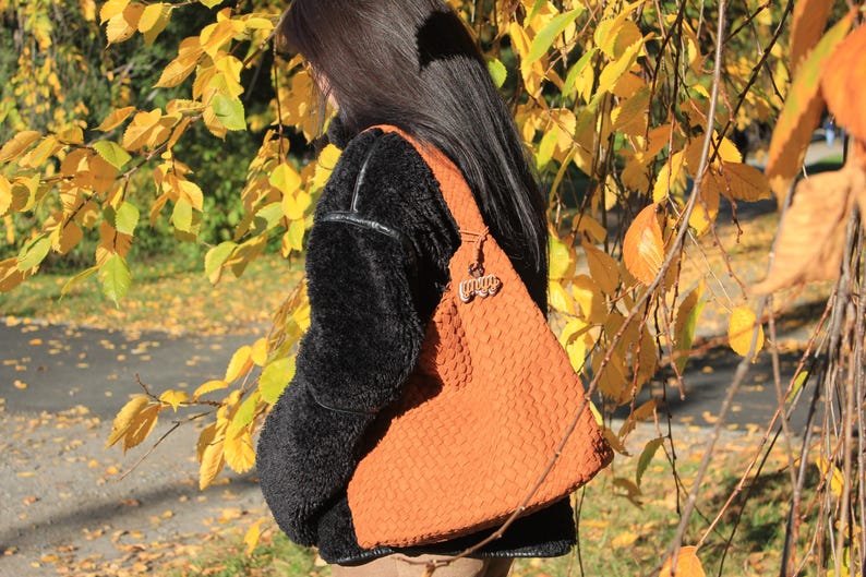 K&ouml;nnte beinhalten: Eine orangefarbene, gewebte Umh&auml;ngetasche mit einem dekorativen Anh&auml;nger. Die Tasche wird &uuml;ber einer schwarzen, flauschigen Jacke getragen. Der Hintergrund zeigt gelbe und gr&uuml;ne Bl&auml;tter, was auf eine herbstliche Umgebung hindeutet.
