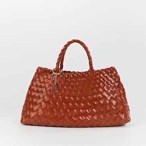 May include: A woven brown leather tote bag with a braided handle and a gold-colored charm. The bag has a trapezoid shape and a textured, basket-weave pattern. The handle is approximately 10 inches tall.