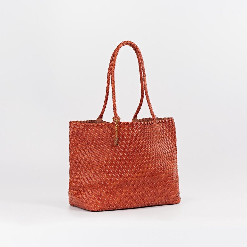 Pode incluir: Uma bolsa tote de couro tecido em tom castanho-avermelhado. A bolsa apresenta al&ccedil;as tran&ccedil;adas e formato retangular. Um pequeno pingente dourado pende da al&ccedil;a. A bolsa &eacute; projetada para transportar itens.