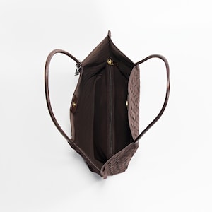 May include: A brown tote bag with woven detailing. The bag has two curved handles and a zippered main compartment. The interior is a darker brown, and the bag appears to be made of leather or a similar material. The bag is empty.
