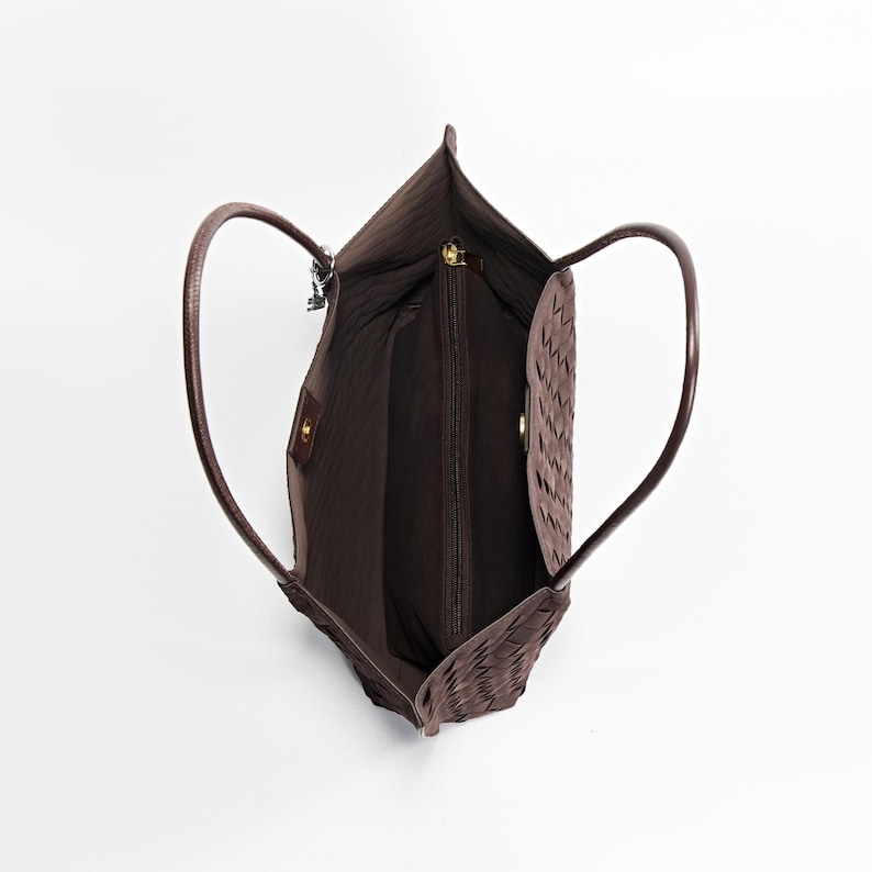 May include: A brown tote bag with woven detailing. The bag has two curved handles and a zippered main compartment. The interior is a darker brown, and the bag appears to be made of leather or a similar material. The bag is empty.