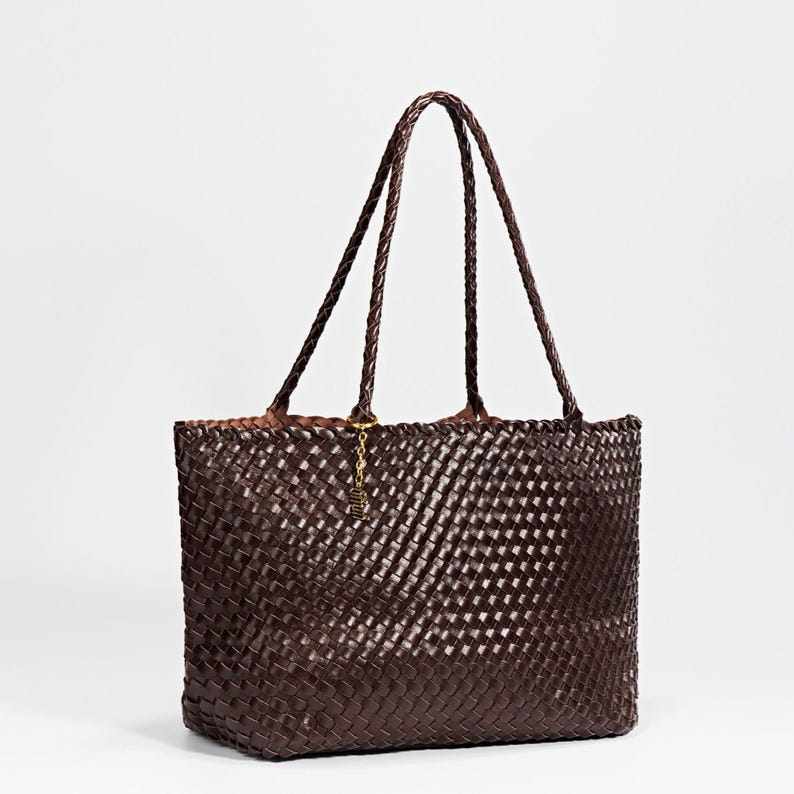 Pode incluir: Uma bolsa tote tecida marrom escuro com al&ccedil;as tran&ccedil;adas. A bolsa tem formato retangular e um padr&atilde;o tecido. Um pingente dourado pende de uma das al&ccedil;as. A bolsa &eacute; feita de um material dur&aacute;vel e &eacute; adequada para uso di&aacute;rio.