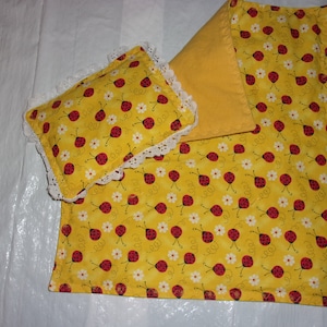 May include: A yellow and white fabric bedding set with a ladybug print. The set includes a pillow with white lace trim and a blanket.