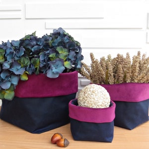 May include: Three fabric storage baskets with burgundy and navy blue fabric. The largest basket is filled with blue and green hydrangea flowers. The middle basket is filled with a decorative white orb. The smallest basket is empty.