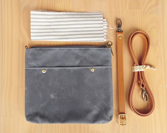 May include: A gray waxed canvas crossbody bag with a brown leather strap and a striped fabric swatch. The bag has a front pocket with two brass rivets and a zipper closure. The leather strap has a brass buckle.