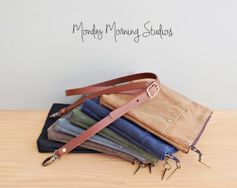 May include: A stack of six canvas bags with a brown leather strap and brass hardware. The bags are in various shades of blue, green, and brown. The strap is folded over the top of the stack and has a brass buckle. The bags are on a wooden surface. The text "Monday Morning Studios" is written in black on a white background above the bags.