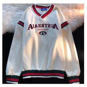 May include: White sweatshirt with a red and green striped collar and cuffs. The sweatshirt has a large embroidered graphic that says "AUSTRALIA" in reverse and a small embroidered graphic of a globe with a kangaroo on it.