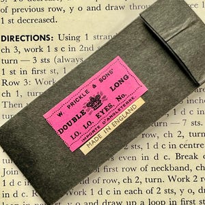 May include: A dark grey paper package with a pink label reading "DOUBLE LO. EYES, No." and "MADE IN ENGLAND." The package is partially open. The background is a page of instructions for a craft project.