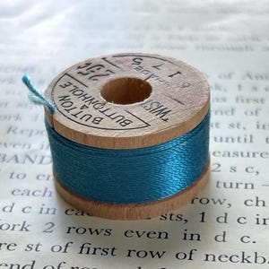 May include: A wooden spool of bright blue thread with a paper label on top that reads BUTTON & BUTTONHOLE, 25 cents, and SHADE 6175. The spool is cylindrical with a central hole and a small length of blue thread extending from the side.