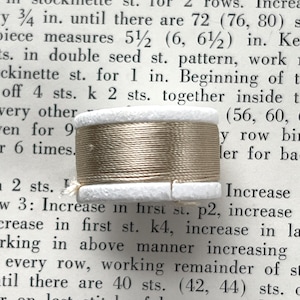 May include: A spool of light brown thread with white cardboard ends rests on a page of printed text. The thread is tightly wound, and the text appears to be instructions for knitting. The spool is a crafting supply.