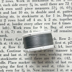 May include: A spool of gray thread with a white cardboard core sits on a page of printed text. The thread is tightly wound around the core, and the text is from a knitting pattern.