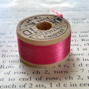 May include: A vintage wooden spool of bright pink pure silk thread. The top label is printed with the text Corticelli Belding, 10 yds, and Pure Silk Twist. The thread is wound around the center of the spool, with a small loose end at the top.