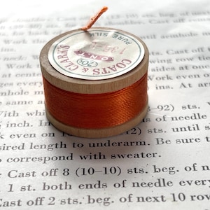 May include: A wooden spool of orange thread with a paper label. The label reads "CLARK'S & COATS" and "PURE SILK". A small piece of thread extends from the top of the spool. The spool is sitting on top of a printed page.