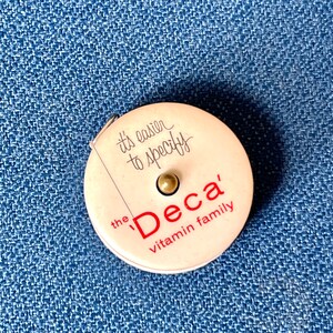 May include: A vintage, round, white tape measure with the text "it's easier to specify" and "the Deca vitamin family" in red. The tape measure has a small metal button in the centre and a measuring tape extending from the side.