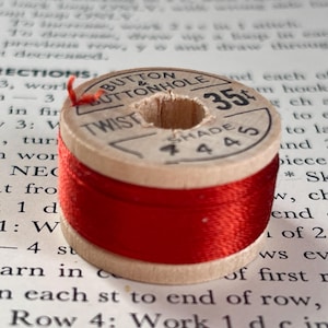 May include: A wooden spool of red thread sits atop a page of printed text. The spool has a label with the words "BUTTON & BUTTONHOLE TWIST" and the price "35¢". The thread is tightly wound around the spool, ready for sewing or crafting.