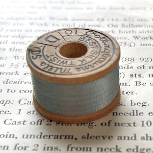 May include: A vintage wooden spool of light blue silk thread. The top of the spool features black printed text that reads "BEST SILK TWIST" and "CORTICELLI MILLS" along with "10 YDS". The spool has a cylindrical shape with a central hole.