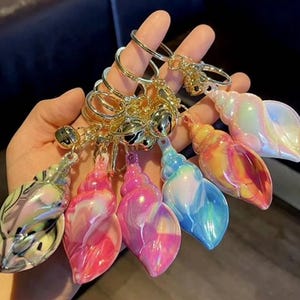 May include: A set of six iridescent seashell keyrings with gold rings and bells. The shells are in various colours, including pink, blue, yellow, and green.