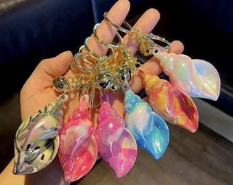 May include: A set of six iridescent seashell keyrings with gold rings and bells. The shells are in various colours, including pink, blue, yellow, and green.
