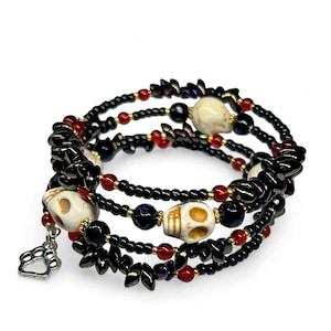 May include: A wrap bracelet featuring black beads, red accents, and cream-colored skull beads. A silver paw print charm hangs from the bracelet. The bracelet is designed to wrap around the wrist multiple times.