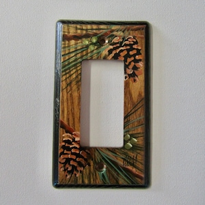 May include: A light switch cover with a painted design of pine cones and needles on a woodgrain background. The cover is black with a dark brown woodgrain pattern.