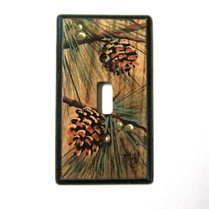 May include: A light switch cover with a painted design of pine branches and pine cones. The background is a woodgrain pattern with a black border.