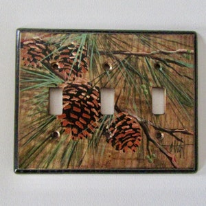 May include: A light brown, green, and black light switch cover with a painted design of pine branches and pine cones. The switch cover has three toggle switches.