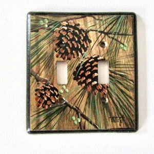 May include: A double light switch cover with a painted design of pine branches and pine cones. The background is a wood grain pattern.