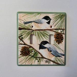 May include: A rectangular double light switch cover plate featuring a hand-painted scene of two black-capped chickadees perched on pine branches with brown pinecones and green needles against a cream background, finished with a sage green border.