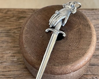 May include: A silver charm in the shape of a hand holding a sword. The charm is detailed and has a ring at the top for attaching to a necklace or bracelet.