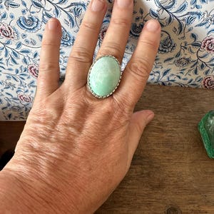 May include: A large oval ring with a light green stone set in a silver bezel. The ring is on a person's finger.