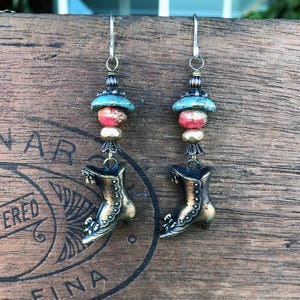 Peut inclure: Une paire de boucles d'oreilles artisanales avec un charme de botte de couleur bronze. Les boucles d'oreilles ont des perles bleues, rouges et dorées. Les boucles d'oreilles sont suspendues à des crochets argentés. Elles sont présentées sur une surface en bois.