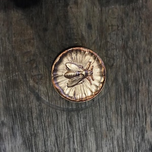 Puede incluir: Un botón redondo de color bronce con un diseño detallado de abeja en el centro. La abeja está rodeada por un patrón de rayos de sol. El botón está sobre una superficie de madera oscura y texturizada.