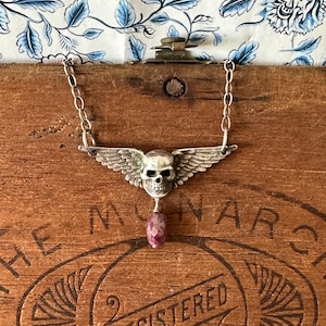 Könnte beinhalten: Silberkette mit einem Anhänger in Form eines geflügelten Schädels und einem rosa Edelstein. Die Kette ist filigran. Die Halskette wird auf einer Holzoberfläche präsentiert. Das Wort "REGISTERED" ist sichtbar.