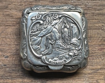 May include: Silver-toned metal box with an ornate design featuring a depiction of the Virgin Mary at Lourdes. The box has a hinged lid and the word "Lourdes" is engraved on the front.