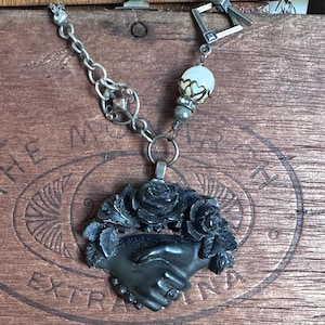 May include: A necklace featuring a black carved pendant of two hands shaking, surrounded by black roses. The chain includes a silver-toned toggle clasp, a round white bead with a metal cap, and a small dark grey faceted bead, connected by silver links.