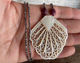 May include: A silver chain necklace with a large, white, intricately carved shell pendant and two dark red gemstone beads.