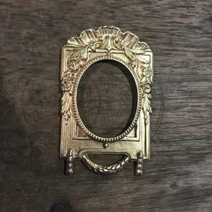 Könnte beinhalten: Ein goldfarbener, verzierter Bilderrahmen mit ovaler Öffnung. Der Rahmen weist florale und blattartige Details, einen dekorativen Griff und eine gewellte Oberseite auf. Der Rahmen ist aus Metall und steht auf zwei kleinen Füßen.