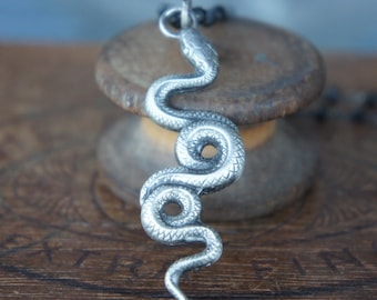 May include: A silver snake pendant necklace with a detailed, textured design. The snake is coiled in a vertical position, with its head at the top and its tail at the bottom.